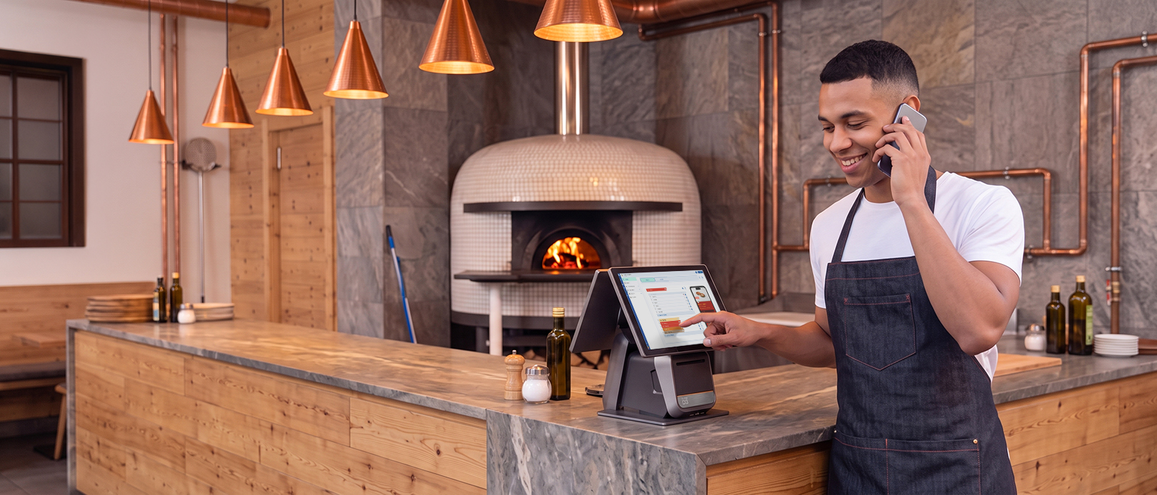 Empleado de una pizzería sonriendo al teléfono mientras utiliza un terminal TPV en el mostrador, con un horno de leña al fondo
