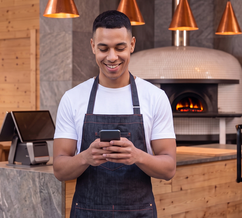 Trabajador sonriente con delantal registrándose en Qromo con el smartphone dentro de una pizzería, con un horno de leña al fondo